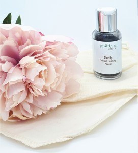 Guiltless Skin's charcoal powder in a glass bottle to the right of the photo. The bottle is standing on an organic cloth next to a pink flower.