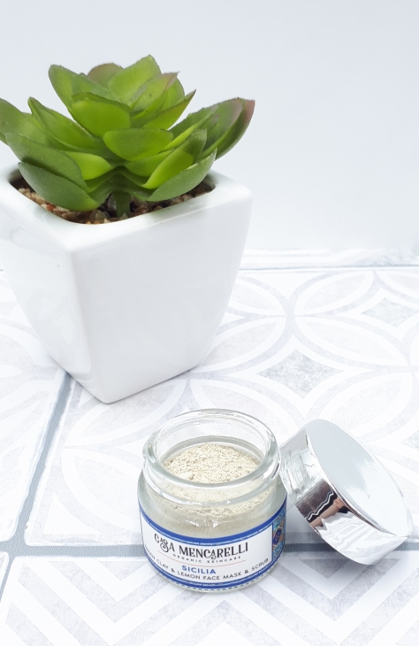 A small glass jar with no lid stands on grey and white Moroccan tiles. The jar contains the clay mask powder which is off-white colour and the lid is propped up against the right hand side of the jar. In the background there's also a small green plant in a white pot.