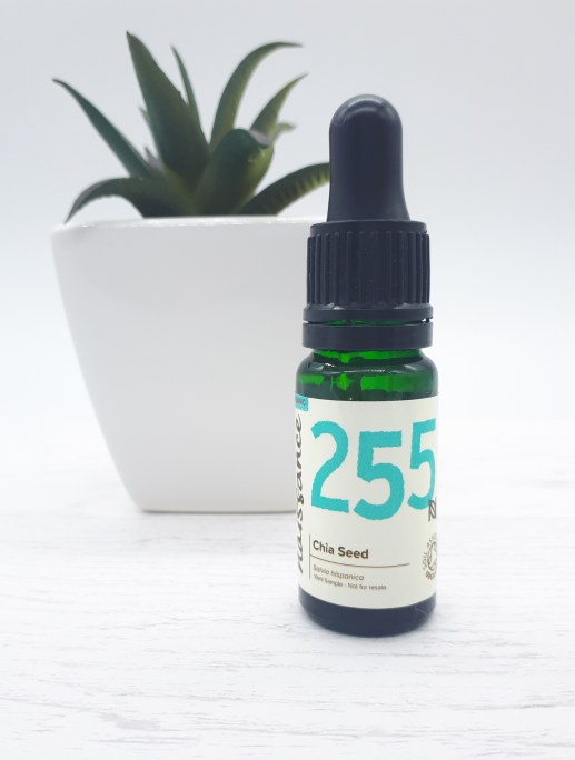 A dark green dropper bottle with a cream label with black and bright blue writing stands on a white wooden floor with white backdrop. In the background, slightly to the left, is a dark green succulent plant with spiky leaves in a white square pot.