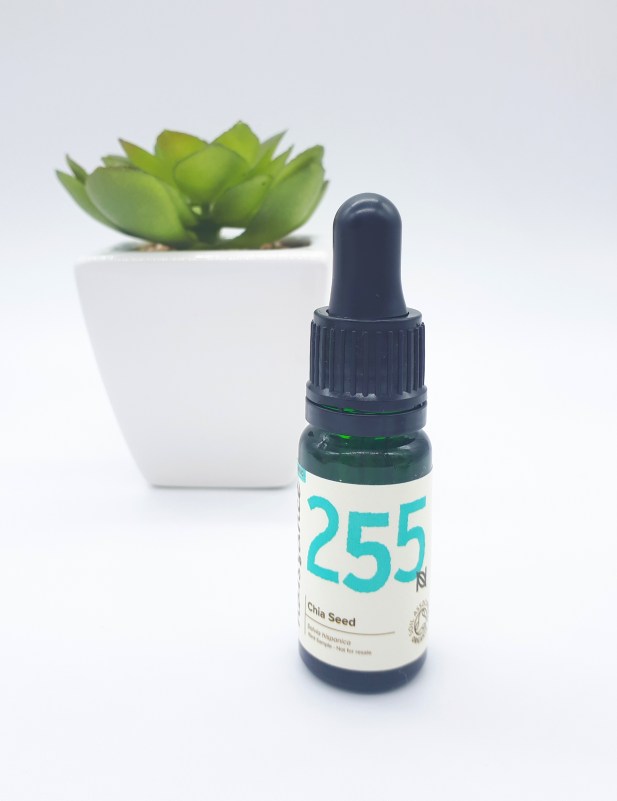A dark green dropper bottle with a cream label with black and bright blue writing stands on a white backdrop. In the background, slightly to the left, is a bright green succulent plant in a white square pot.
