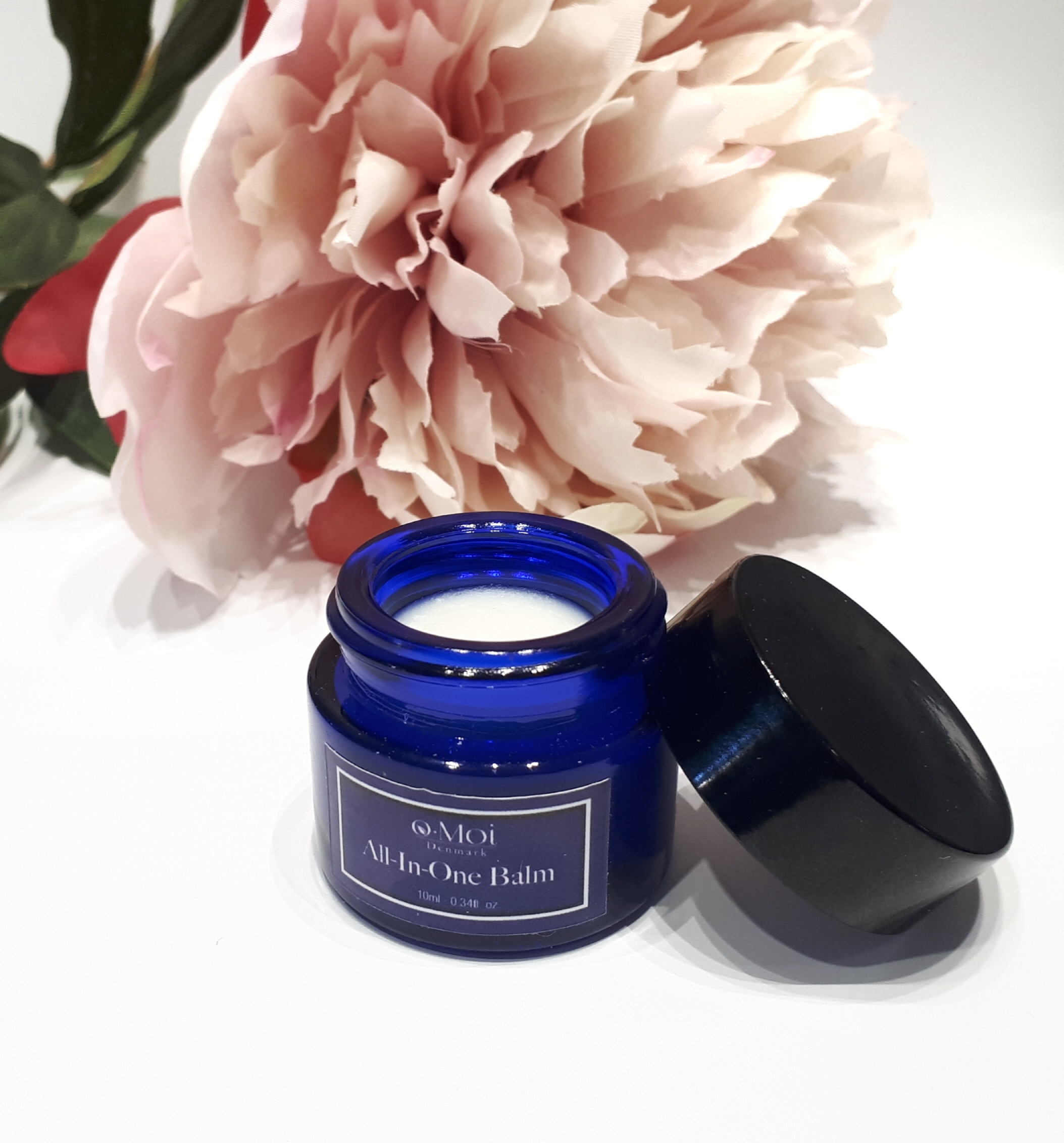 A small blue jar with a dark blue label stands on a white background in front of a pink flower. The lid is leaning up against the jar so the white balm inside is exposed.