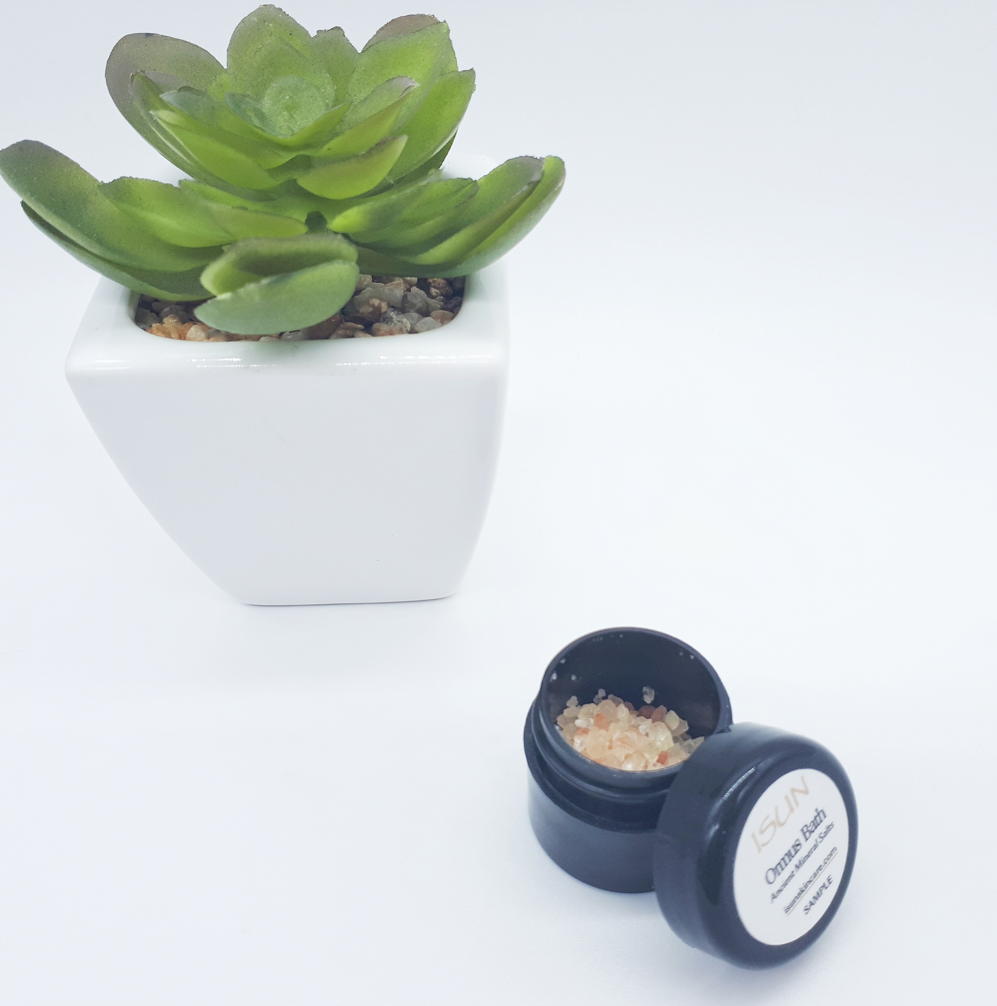 A small black pot containing amber and orange bath salts stands on a white background. On the left, slightly behind the pot is a green plant in a white square pot.