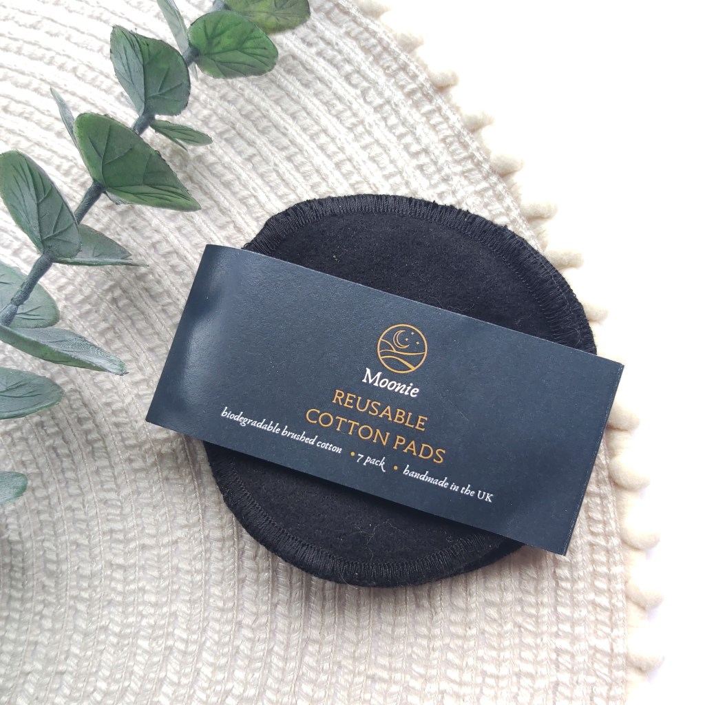 A pack of Moonie Reusable Black cotton pads on a beige place mat with pom pom edge and a eucalyptus stem. A Plastic Free Green Beauty Guide by Beauty Folio highlighting plastic pollution and plastic free beauty from Lovethical. 