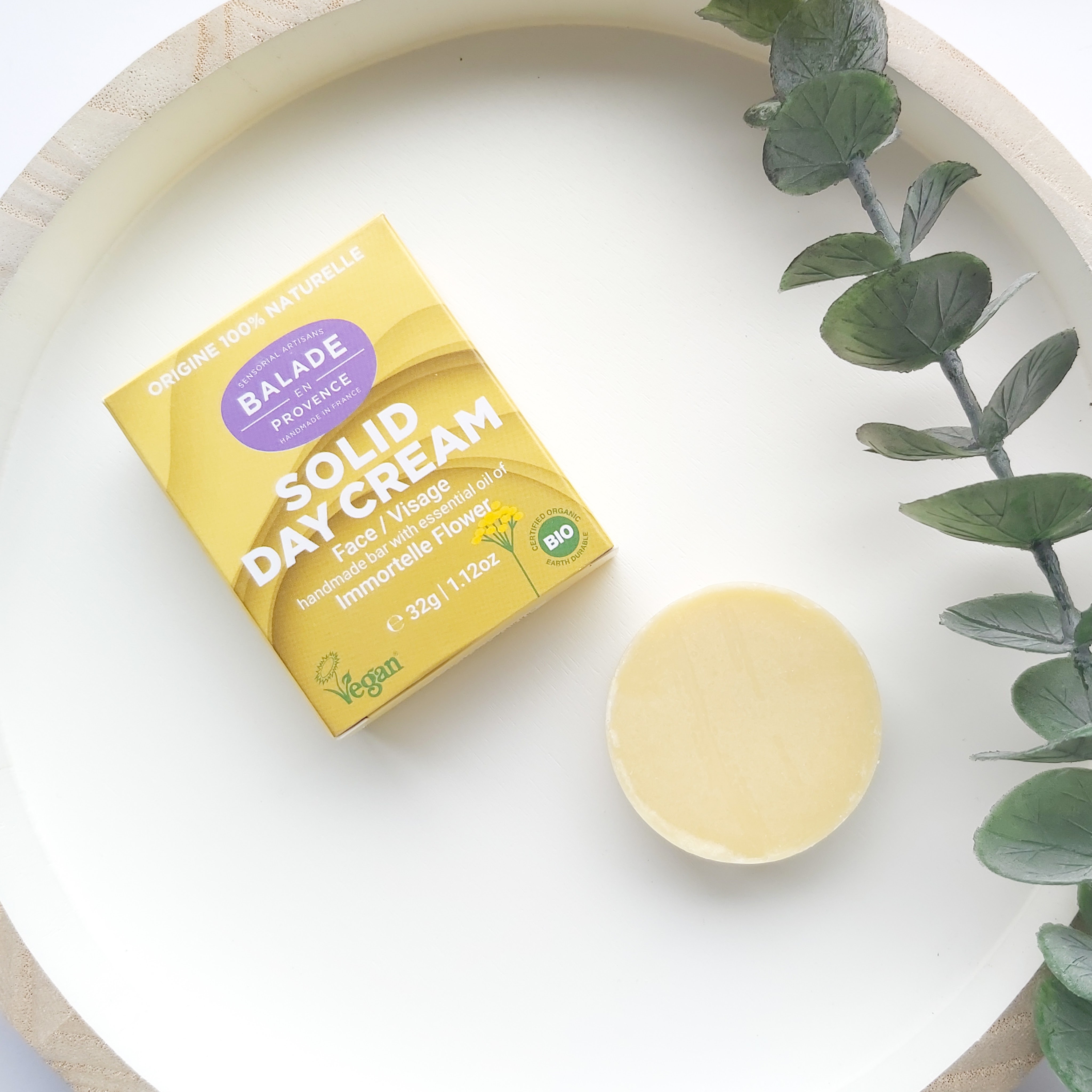 A round, yellow-coloured solid day cream bar by Balade En Provence sits on a white, wooden round tray with a eucalyptus stem to the side and yellow, branded outer box just above - Freedm Street Subscription Beauty Box review by Beauty Folio