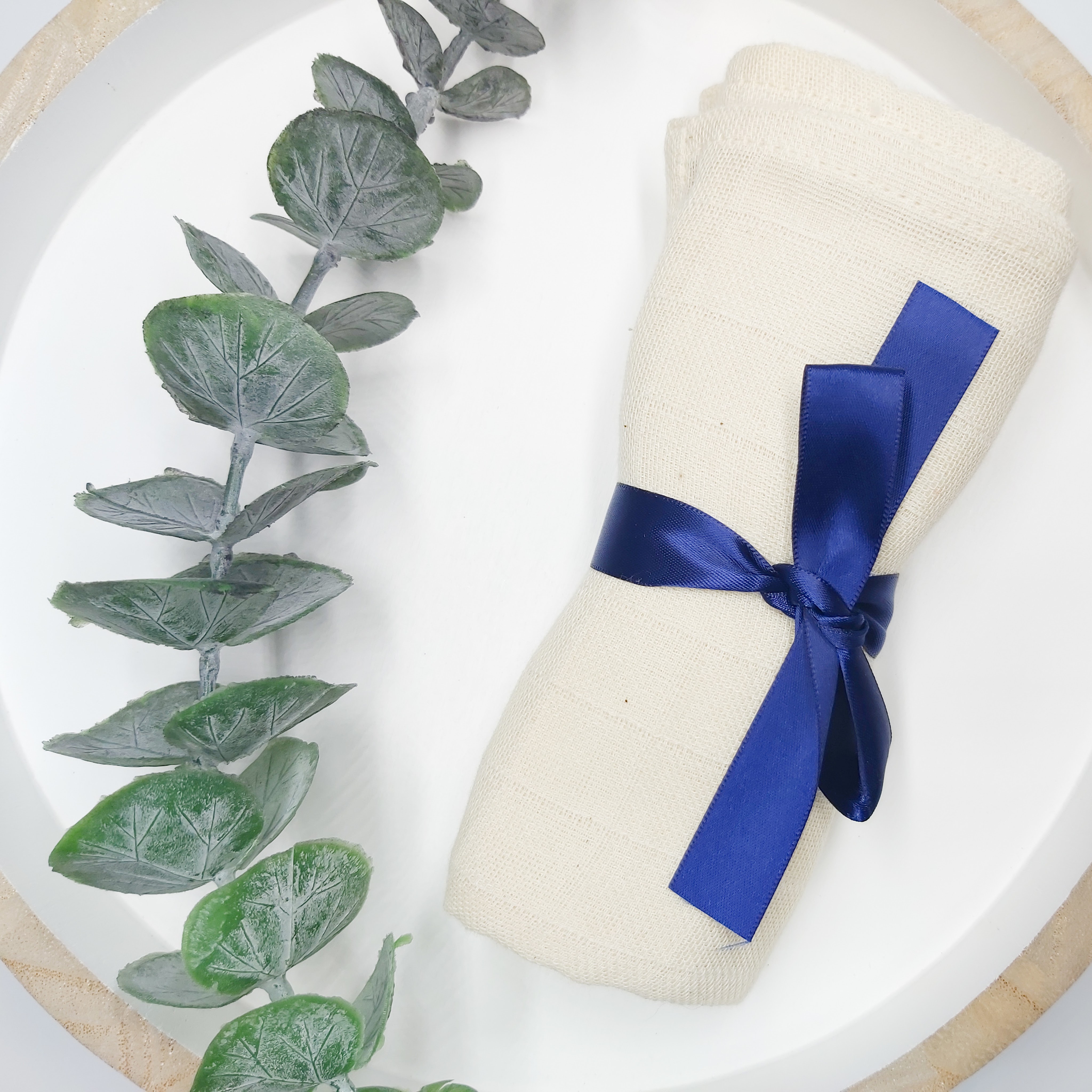 Amaranthine's Muslin Cleansing Cloths tied with a blue ribbon on a white and wood tray with a eucalyptus stem to the side.