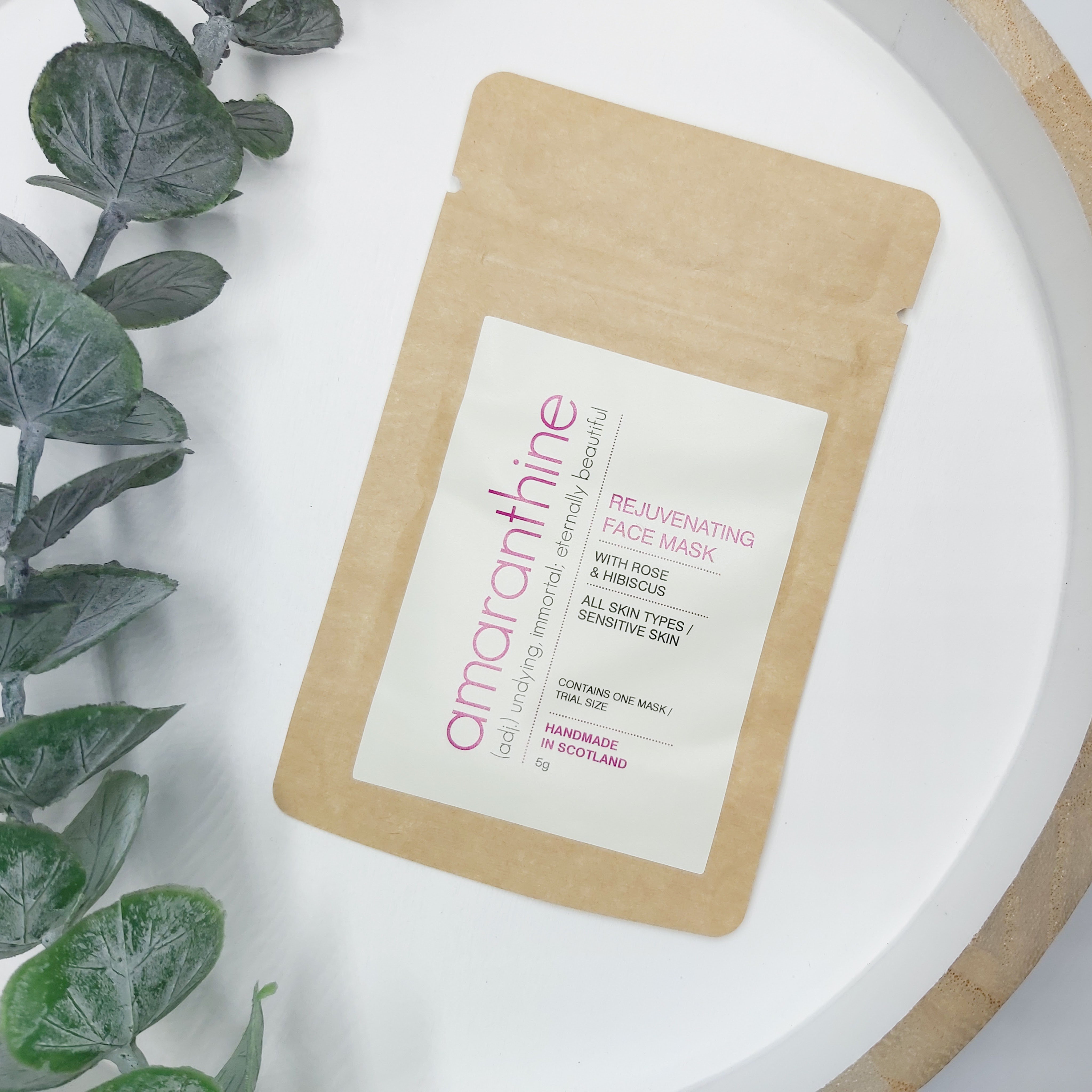 A kraft pouch of Amaranthine's Rejuvenating Face Mask on a white and wood tray with a eucalyptus stem to the side.