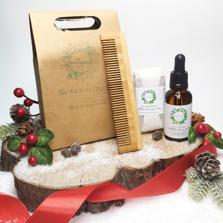 Shampoo bar, hair oil and bamboo come standing next to cardboard gift box on a log slice with Christmas foliage and red ribbon. Natural Beauty Christmas Gift Guide by Beauty Folio 