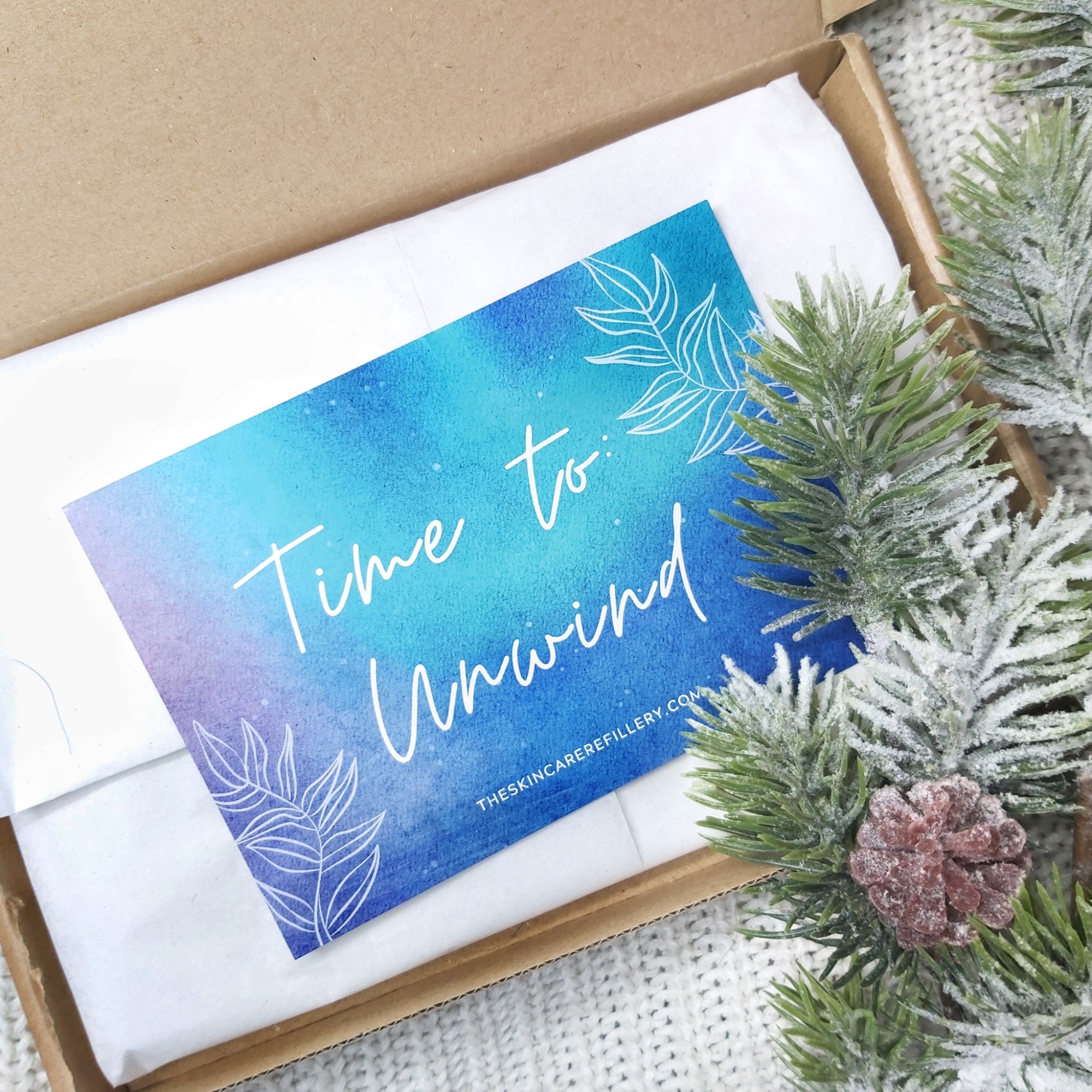 An open letterbox gift box from The Skincare Refillery with a Time to Unwind blue information card. Knitted blanket background with Christmas foliage to the corner. Natural Beauty Christmas Gift Guide by Beauty Folio