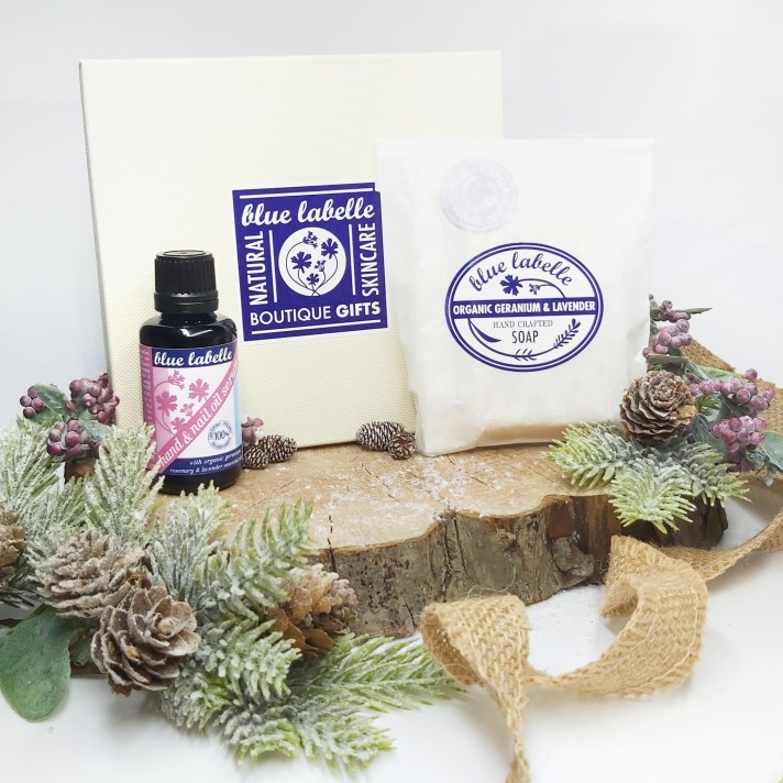 The Blue Labelle branded gift box with serum and soap standing on a log slice with Christmas foliage and jute ribbon. Natural Beauty Gift Guide by Beauty Folio