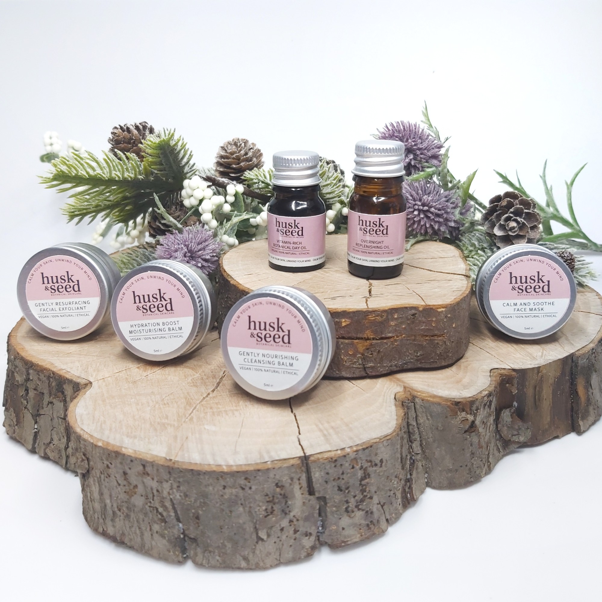 Husk & Seed Skincare Facial Collection Sample Set miniature bottles and tins sat on 2 log slices with Christmas foliage. Natural Beauty Christmas Gift Guide by Beauty Folio