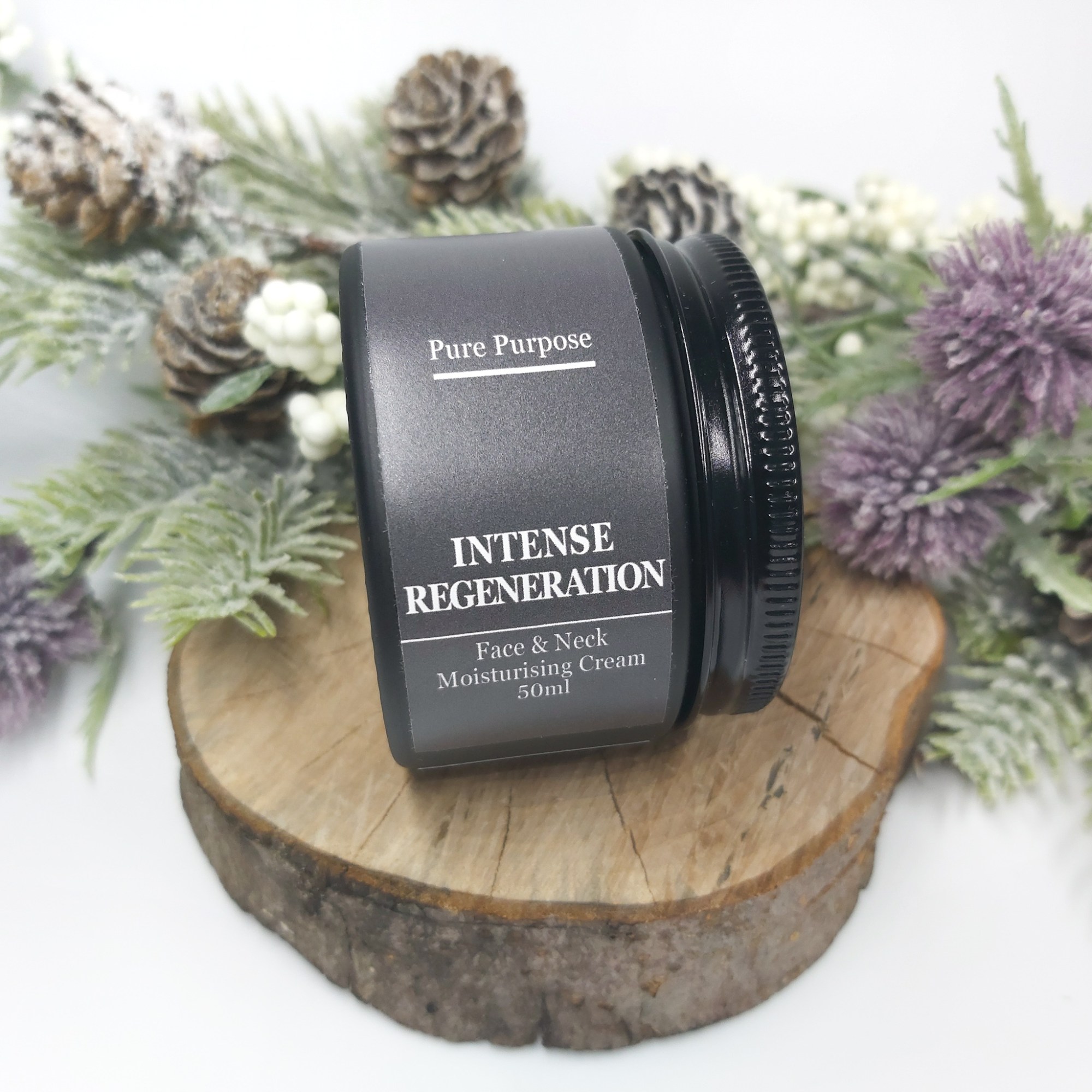 Pure Purpose Intense Regeneration Face & Neck Cream in a black jar is sat on a log slice with Christmas foliage. Natural Beauty Christmas Gift Guide by Beauty Folio