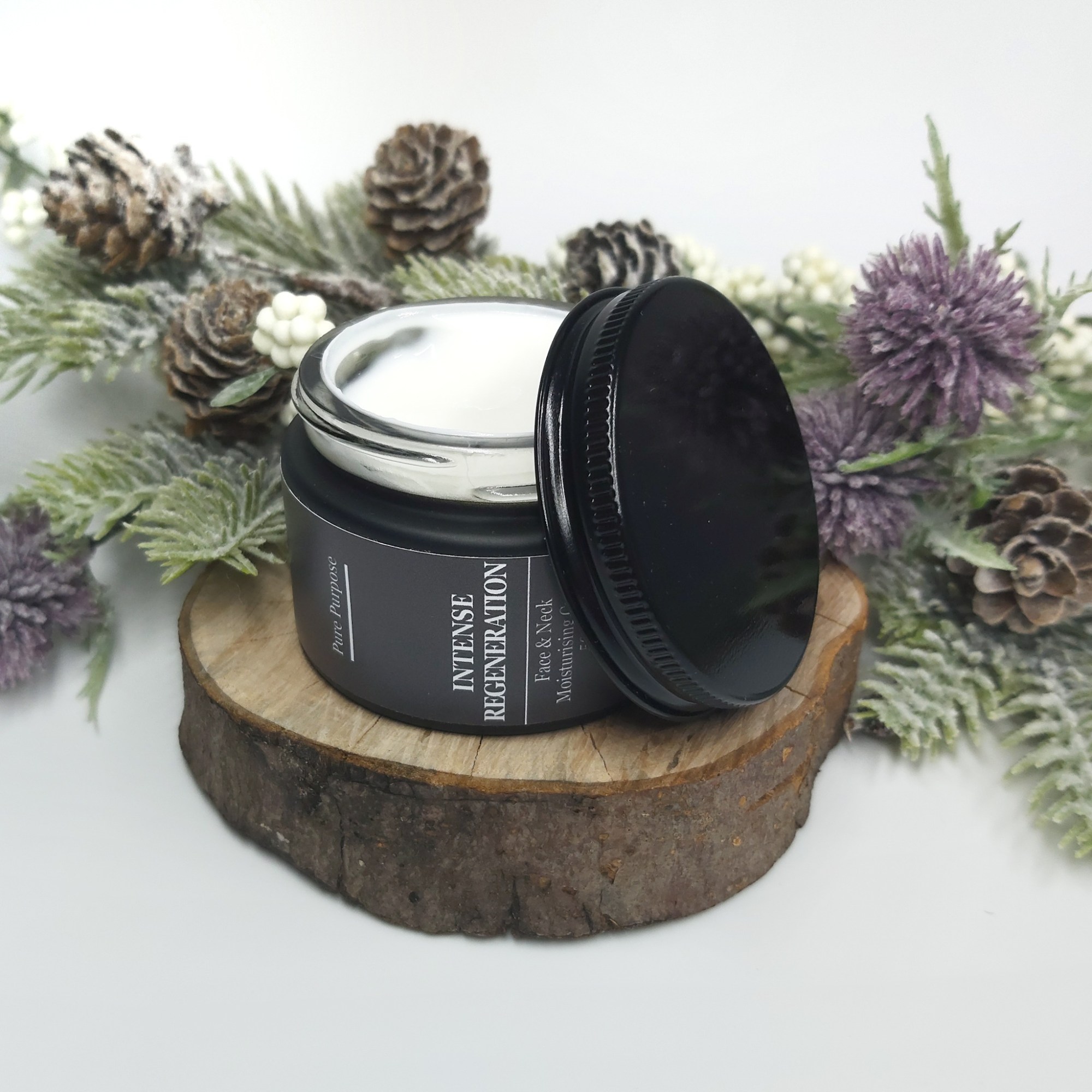Pure Purpose Intense Regeneration Face & Neck Cream in a black jar without the lid is sat on a log slice with Christmas foliage. Natural Beauty Christmas Gift Guide by Beauty Folio