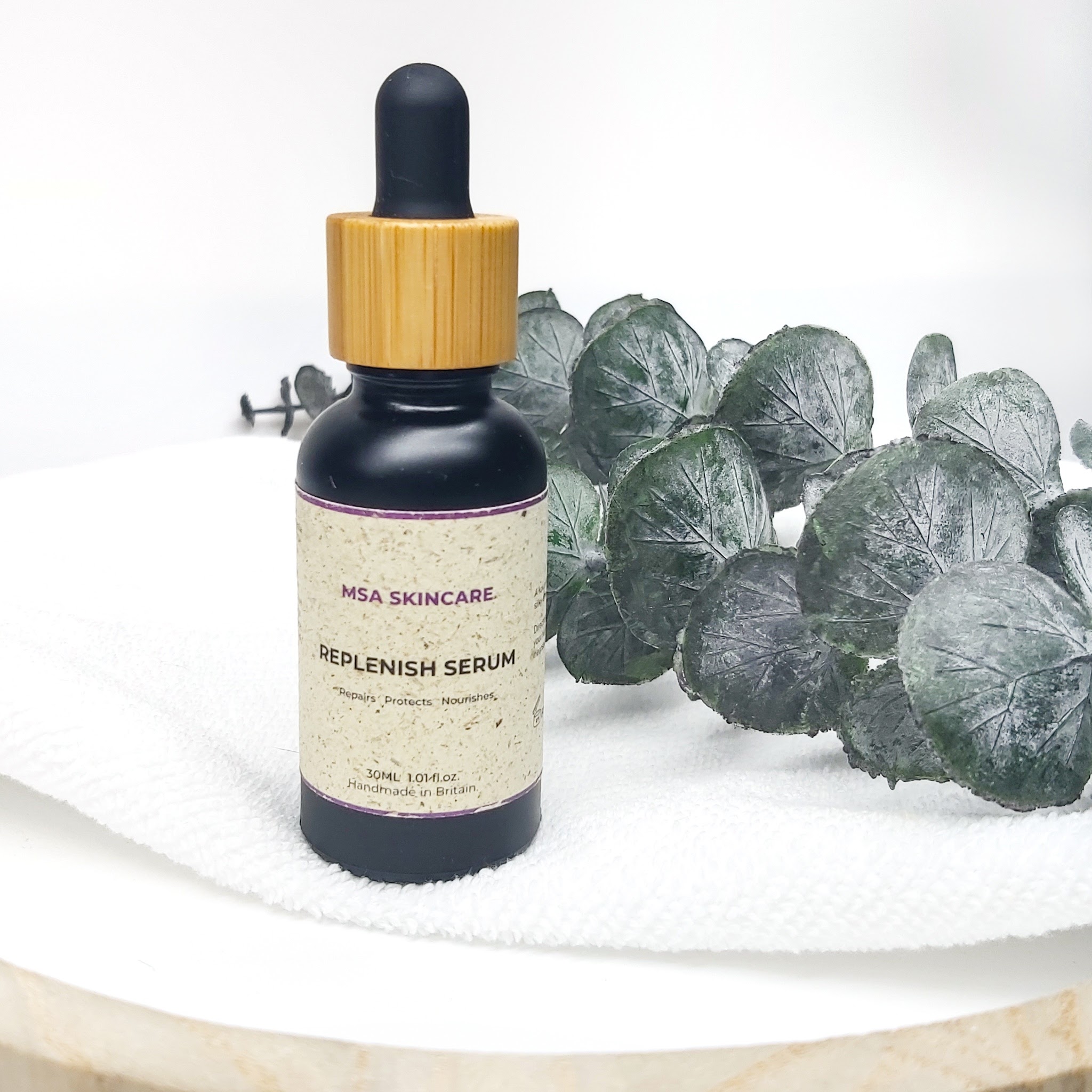MSA Skincare Replenish serum on a white towel, which is on a white and wood round tray with a stem of eucalyptus stem in the background. Natural skincare review by Beauty Folio