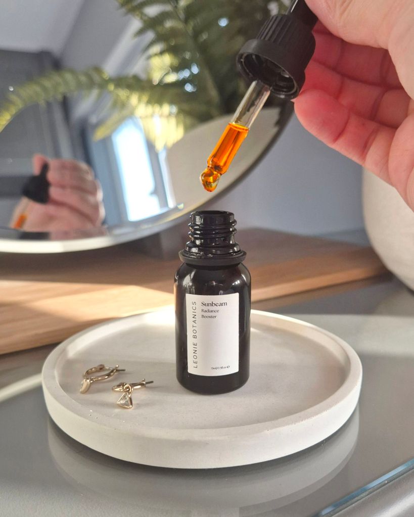 Sunbeam dropper shot in front of a round dressing table mirror. The black bottle is sat on a white concrete dish with earrings next to it - Photos by Beauty Folio