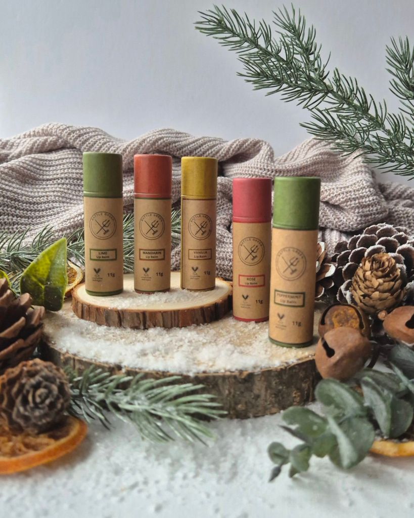 Battle Green Lip Balms on a log slice with foliage and artificial snow - Photo by Beauty Folio, Green Beauty Content Creator