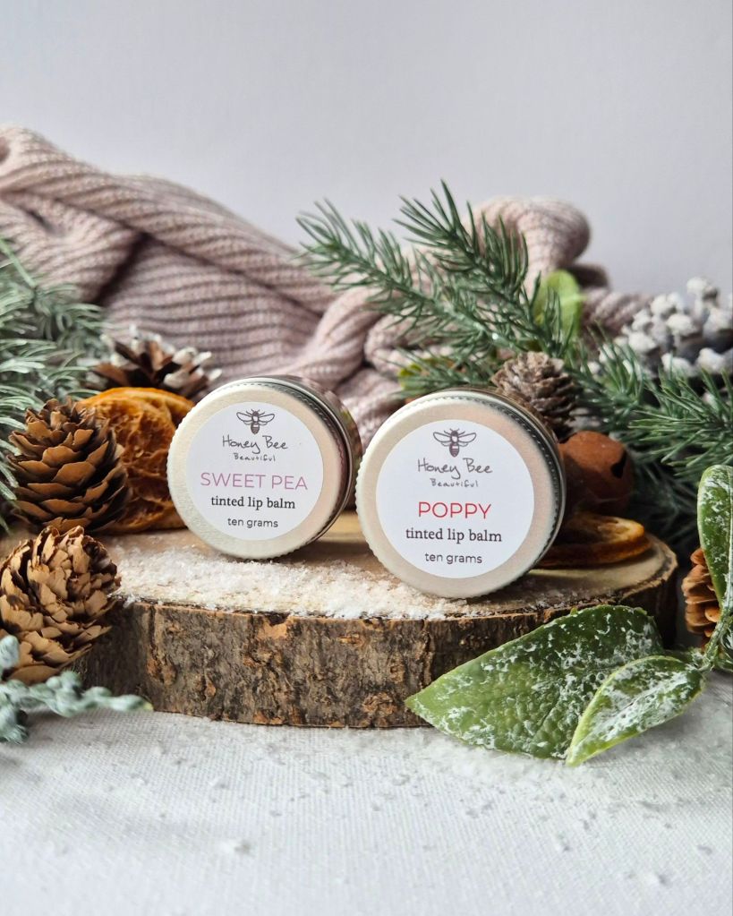 Two honey Bee Beautiful jars on a log slice with foliage and artificial snow - Photo by Beauty Folio, Green Beauty Content Creator