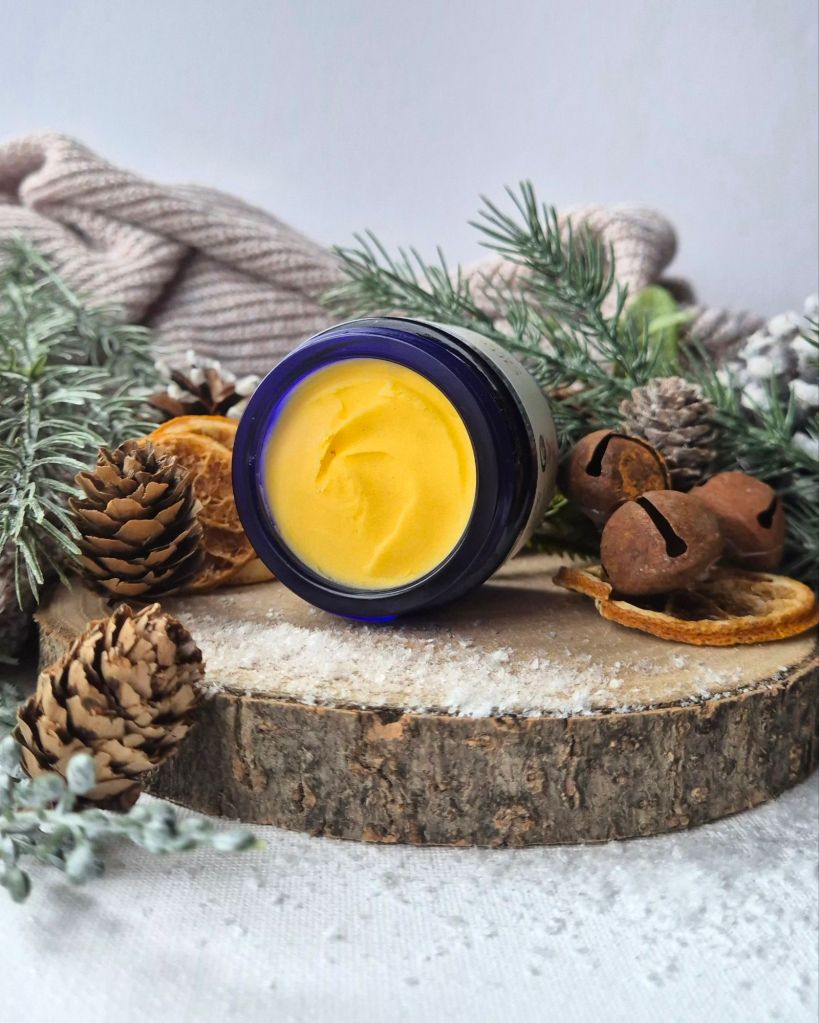 Amaranthine's Body Creme on it's side, open, on a log slice with foliage and artificial snow - Photo by Beauty Folio, Green Beauty Content Creator