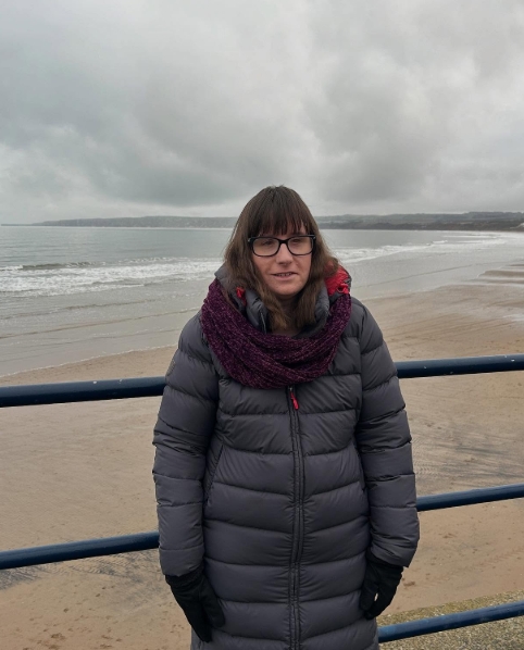  Holly standing in front of a metal railing on a promenade by the beach. She’s wearing a long, puffa navy coat, black gloves, and a burgundy scarf. She has long brown hair and is wearing glasses. The background features a sandy beach and the sea, with waves gently lapping at the shore. The sky is grey and moody.]