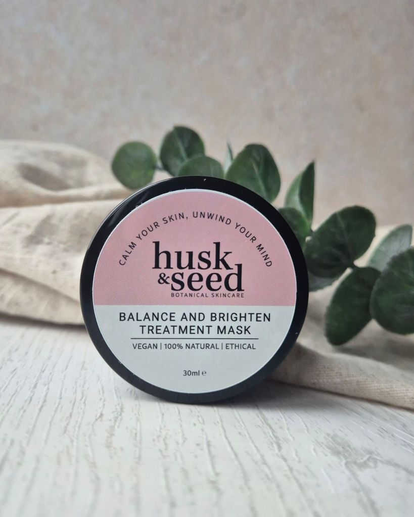 Jar of husk & seed Balance and Brighten Treatment Mask on a wooden surface, surrounded by fabric and greenery.