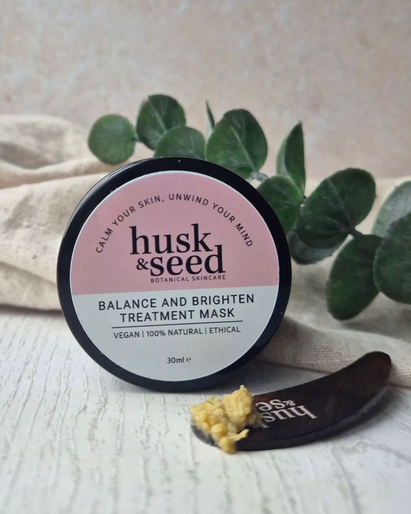 A jar of 'husk & seed' Balance and Brighten Treatment Mask, 30ml, displayed on a wooden surface with a small wooden spoon holding a sample of the mask. Surrounding elements include green foliage and a textured fabric.