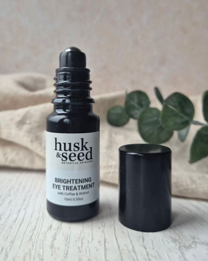 A bottle of husk & seed Brightening Eye Treatment with a rollerball applicator, placed next to its black cap on a textured surface. The background features a soft fabric and greenery.